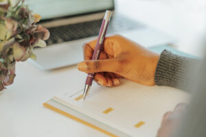 Pictured is a black woman's hand holding a purple The Circle pen and is writing in a yellow More Than Profit Journal