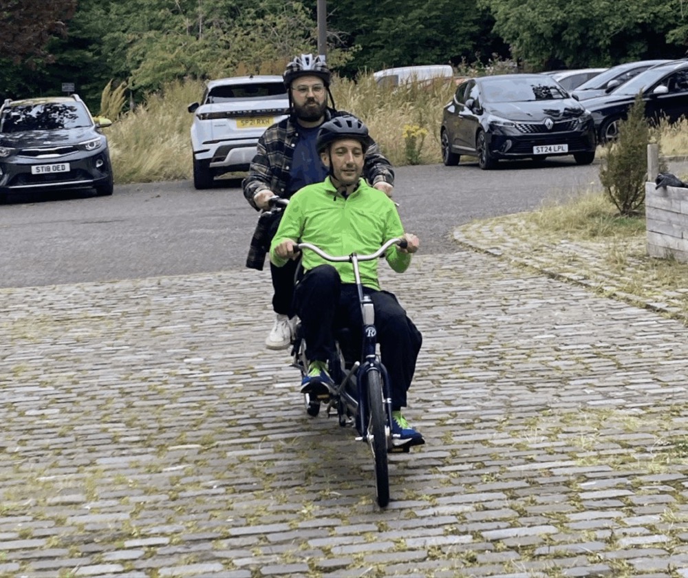 Two men are riding a tandem bike on a cobbled patch of road